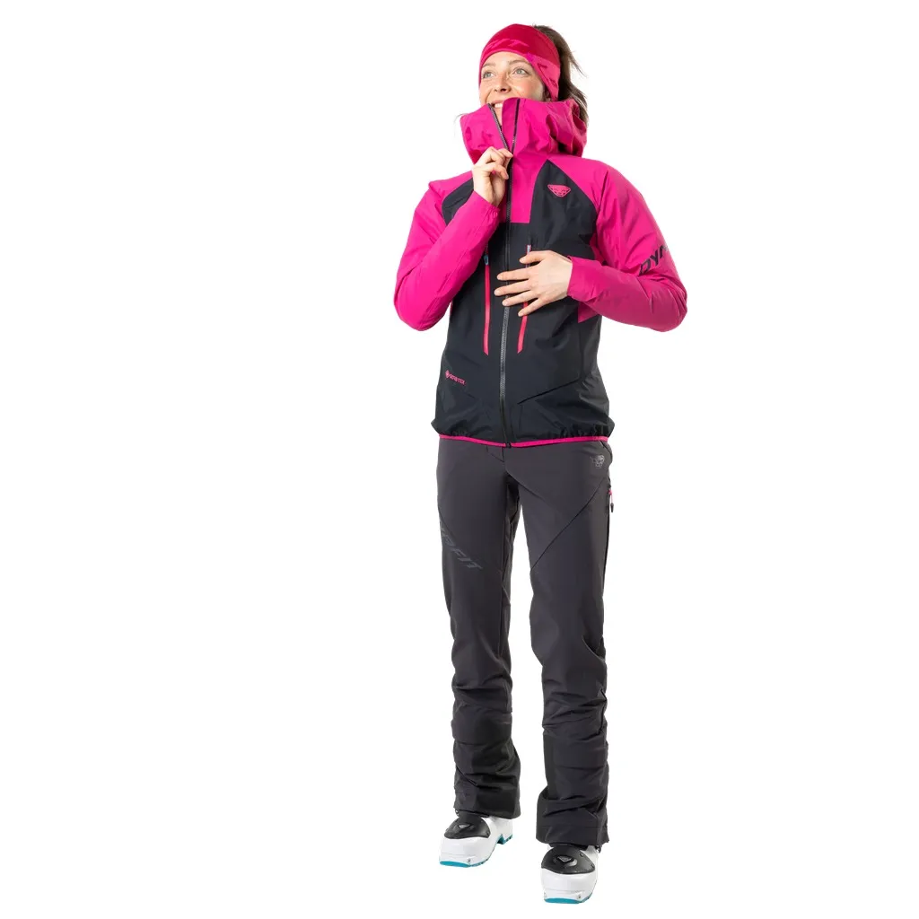 Frau in Dynafit TLT Touring DST Hose, schwarz, pinke Jacke, Skischuhe, Blick nach oben.