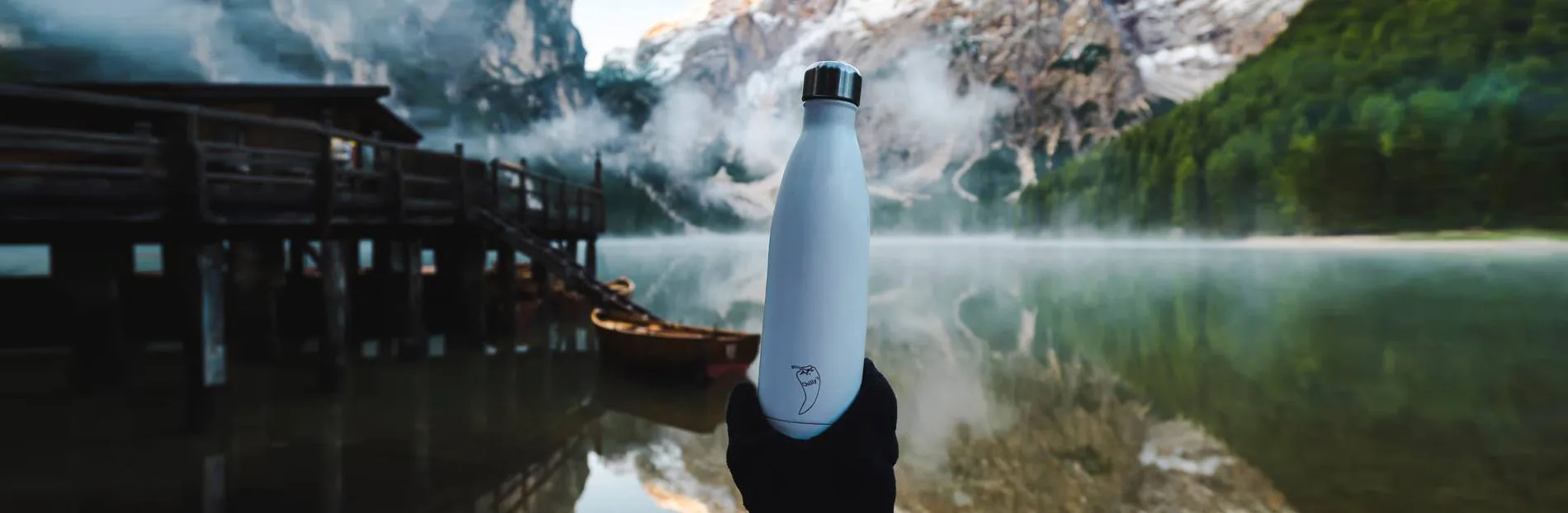 Eine weiße Chilly's Trinkflasche vor einem Bergsee in einer Hand gehalten. Ein Holzsteg und Boote sind im Hintergrund zu sehen.