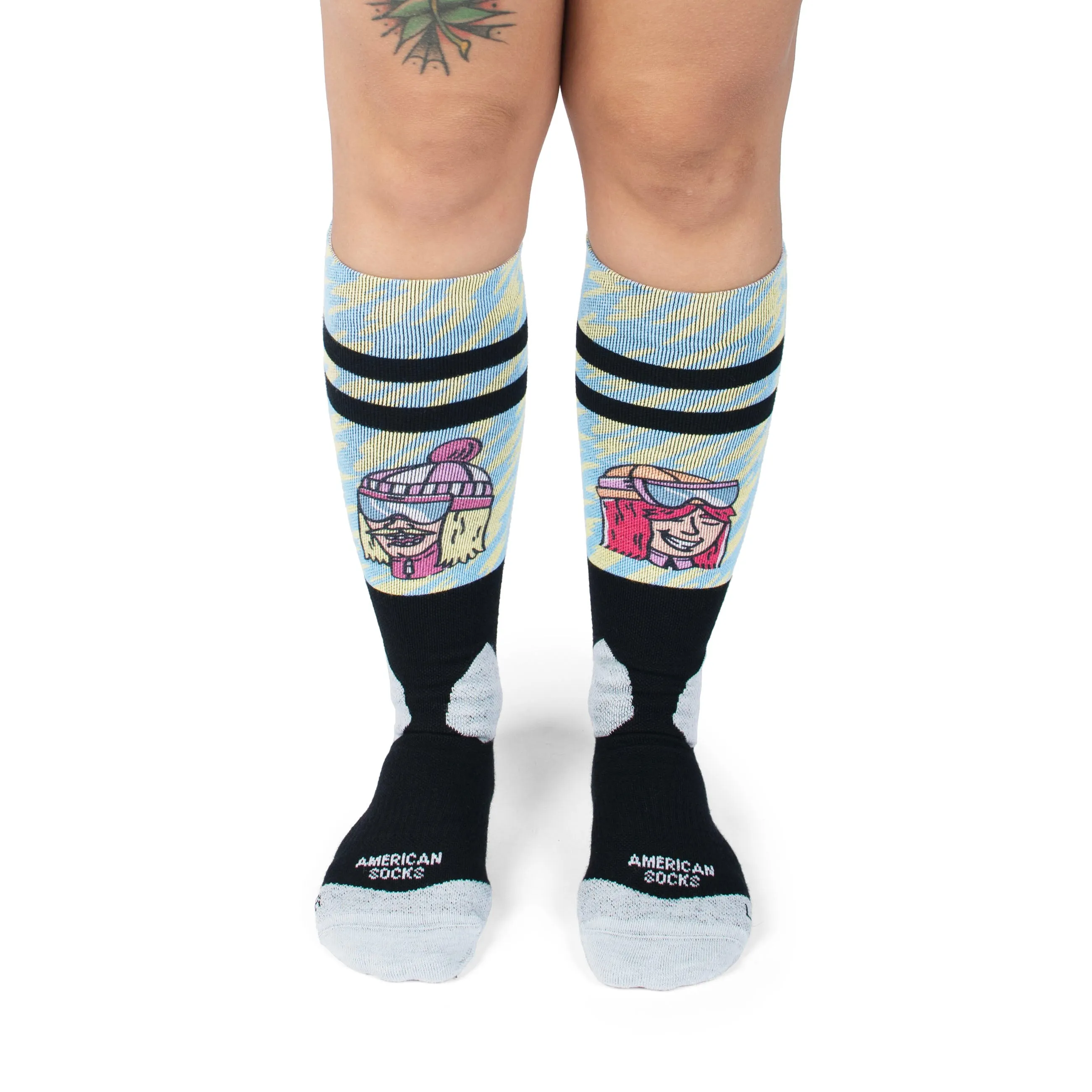 Beine in Slope Dope Snow Socks mit Ski-Motiven, blauen Mustern und schwarzen Streifen. "American Socks" Schriftzug sichtbar.