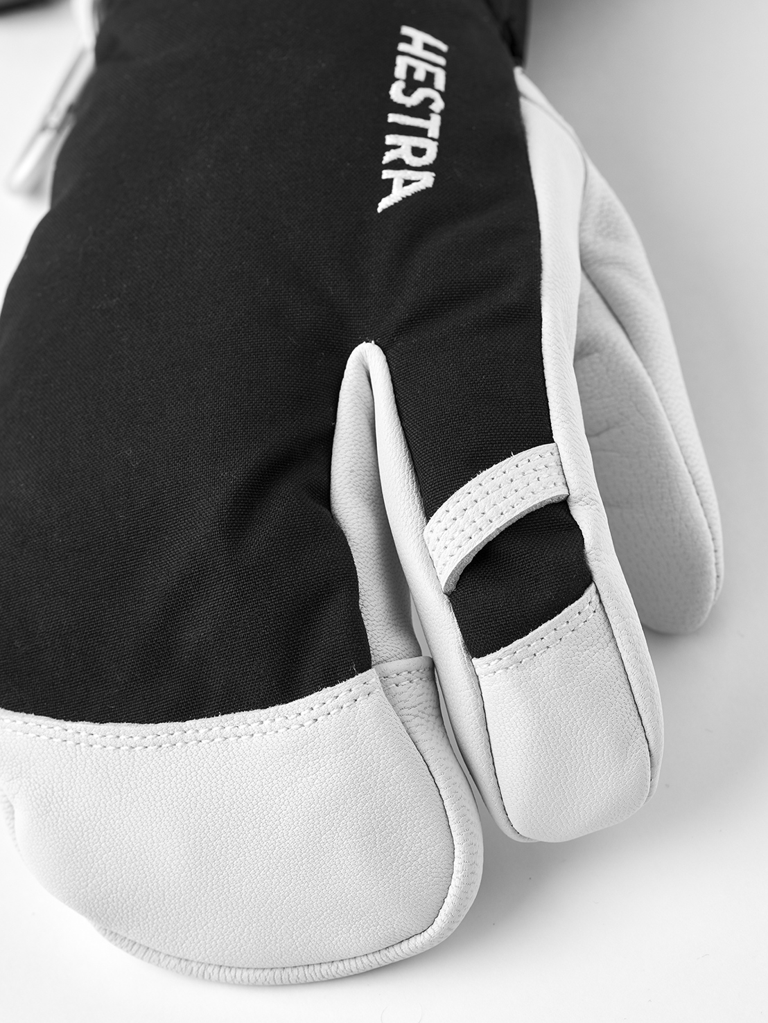 Hestra 3-Finger-Handschuh Nahaufnahme eines schwarzen Hestra 3-Finger-Handschuhs mit weißem Leder an Fingern und Daumen. Das Hestra Logo ist vertikal zu sehen.