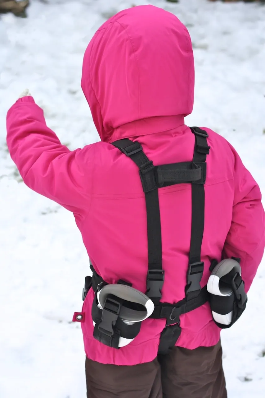 Kind mit rosa Jacke und Lil' Ripper Gripper Skilernhilfe am Rücken im Schnee.
