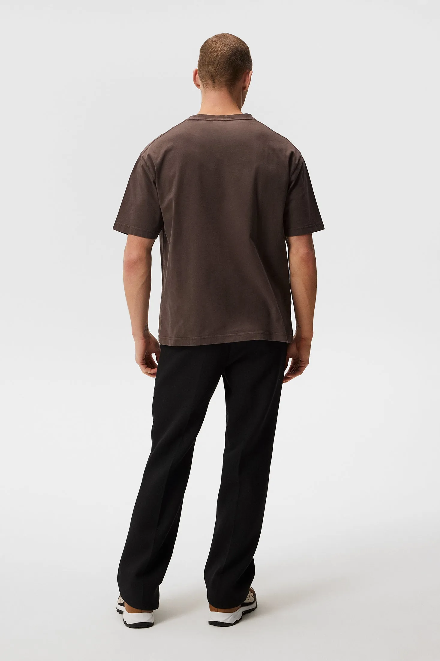 Ein Mann von hinten, trägt ein braunes T-Shirt und schwarze Hose vor einem weißen Hintergrund.