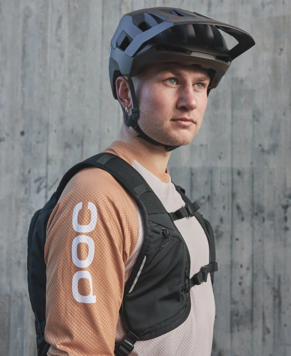 Ein Mann mit Fahrradhelm und Column VPD Backpack 13L vor einer Betonwand. Er trägt ein langärmliges Hemd mit "POC" auf dem Ärmel.