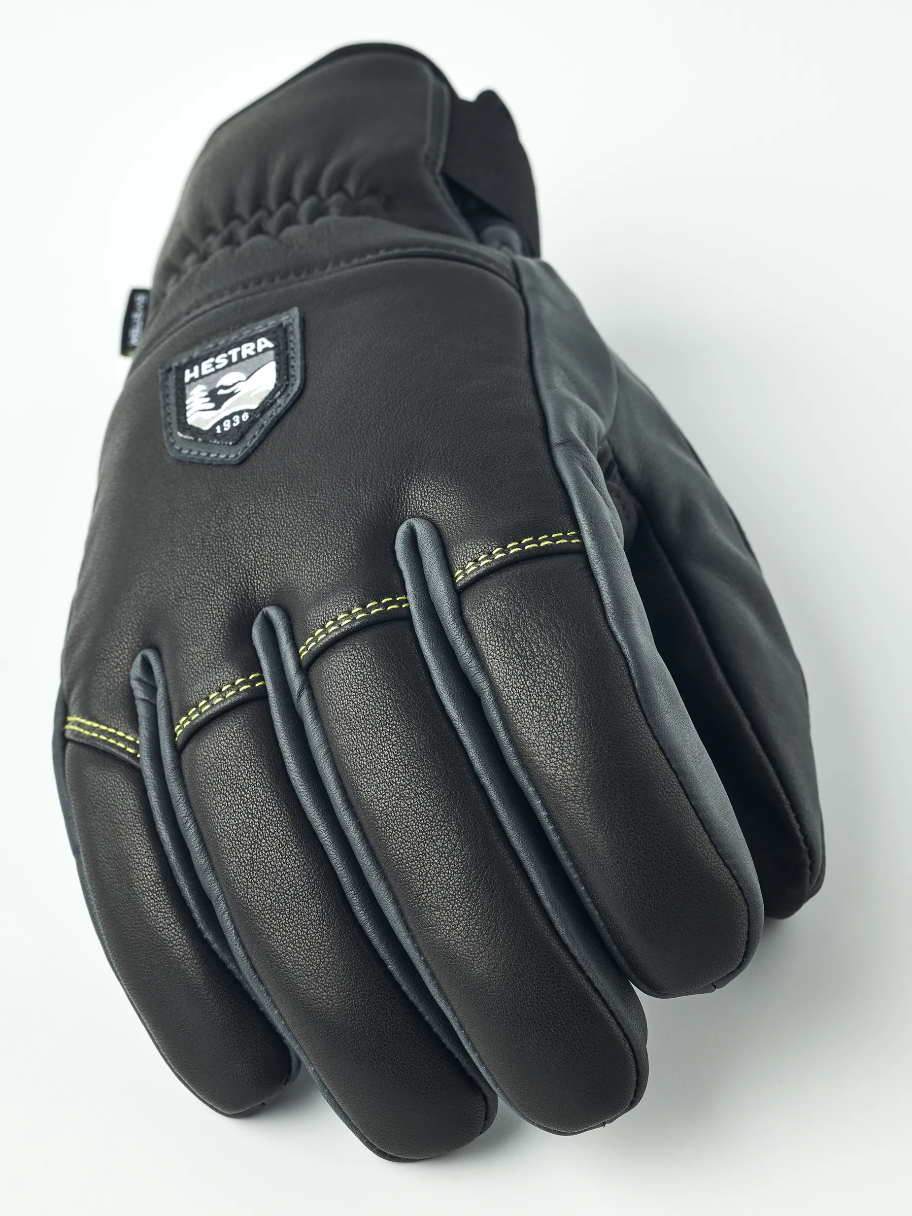 Detailansicht des schwarzen Hestra Omni Trigger Handschuhs aus Leder mit Logo-Aufnäher und gelber Naht.