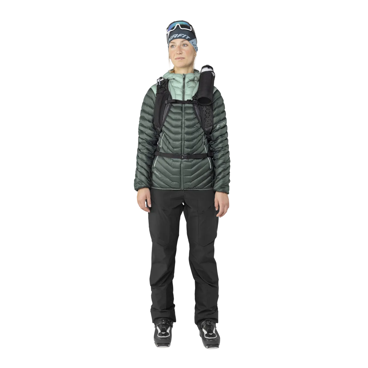 Eine Frau in Ski-Bekleidung, mit Dynafit Ridge Ultralight Daunenjacke in Grün, Hose und Skischuhen.