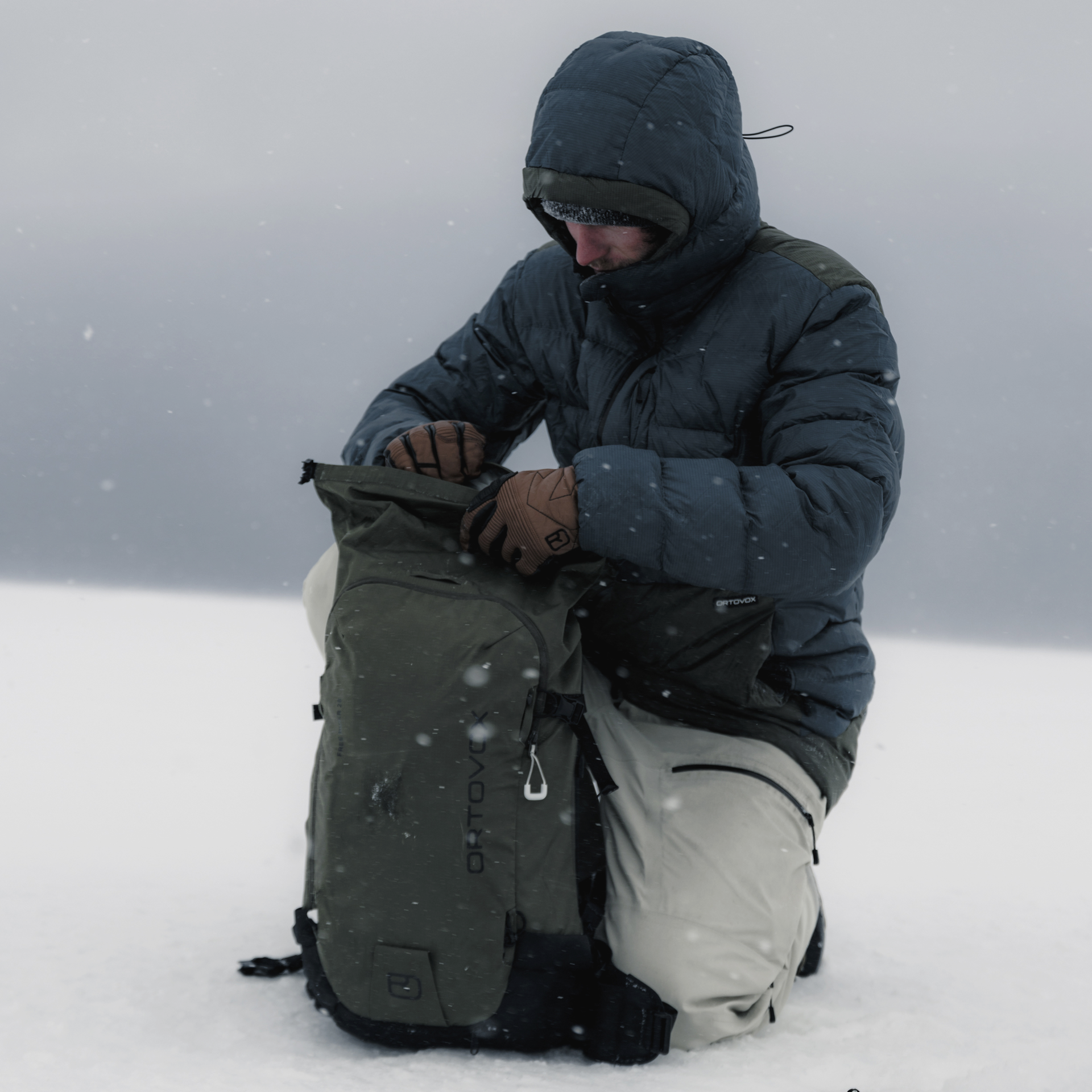 Mann im Schnee mit Rucksack Ein Mann kniet im Schnee, er trägt eine Daunenjacke und öffnet einen olivgrünen Rucksack. Es schneit leicht.