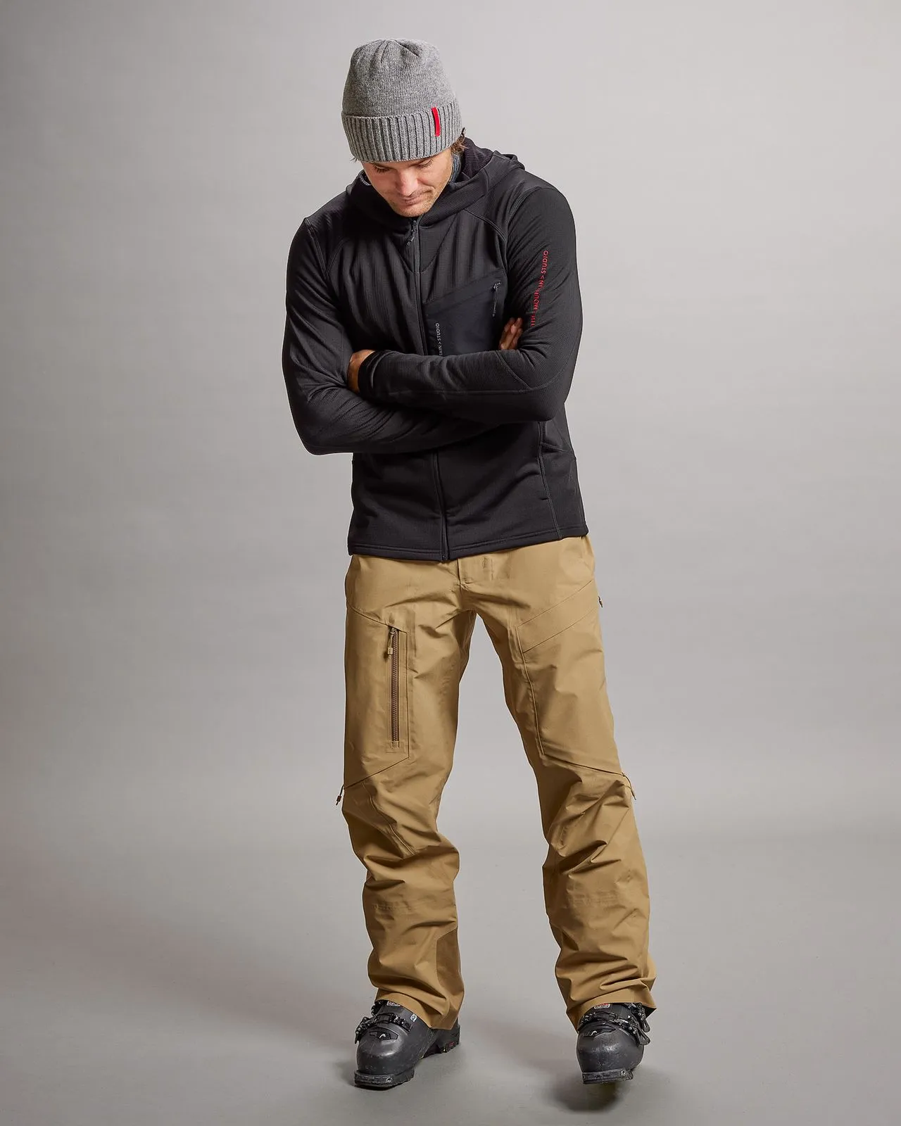 Mann mit schwarzer M-4 Techno Stretch Mid Hood, beiger Hose und grauer Mütze, posiert mit verschränkten Armen.