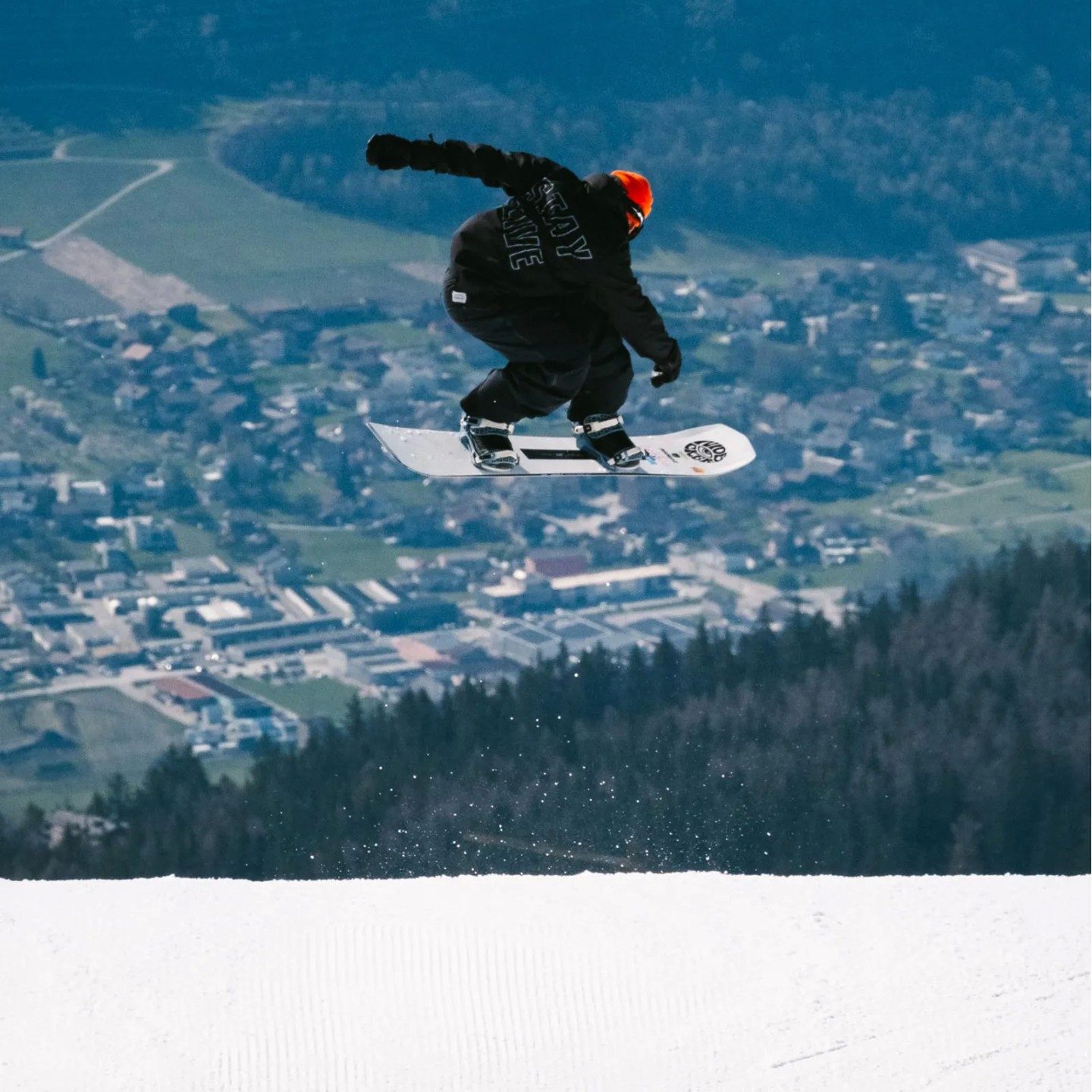 Snowboarder mit oranger Mütze springt auf dem Nidecker Thruster Snowboard über eine präparierte Piste.