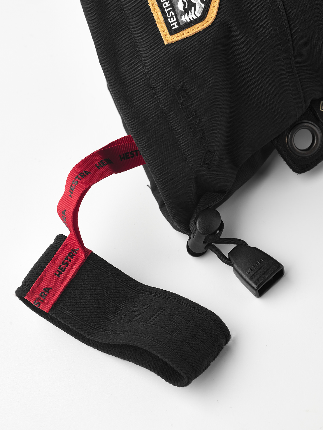 Schwarze Hestra Fäustlinge Nahaufnahme eines schwarzen Hestra Fäustlings mit Gore-Tex, roter Schlaufe und Logo-Patch. Details wie Kordelzug und Öse sind sichtbar.