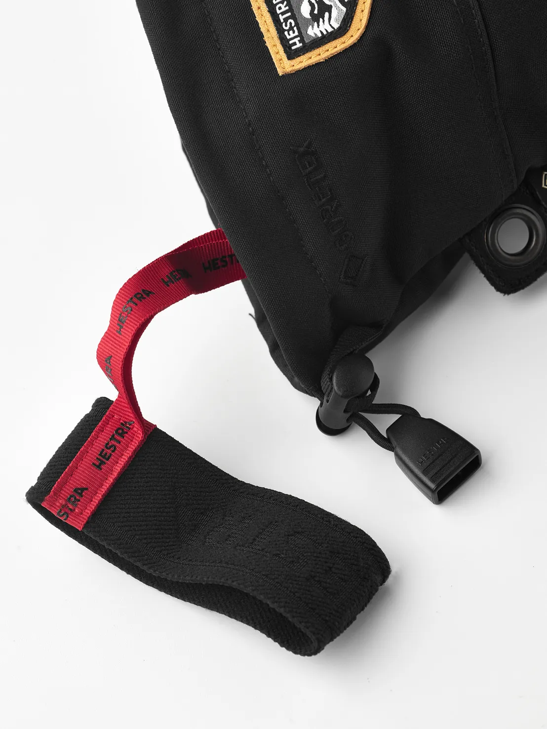 Nahaufnahme eines schwarzen Hestra Fäustlings mit Gore-Tex, roter Schlaufe und Logo-Patch. Details wie Kordelzug und Öse sind sichtbar.