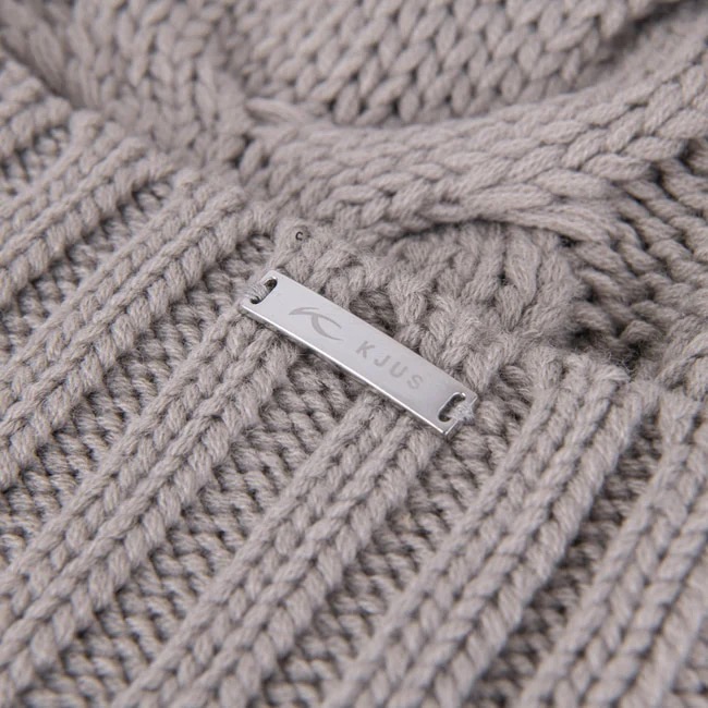 Nahaufnahme einer hellgrauen Strickmütze mit Zopfmuster und einem silbernen "KJUS"-Logo-Etikett.