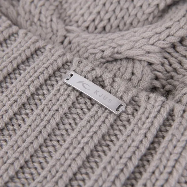 Nahaufnahme einer hellgrauen Strickmütze mit Zopfmuster und einem silbernen "KJUS"-Logo-Etikett.