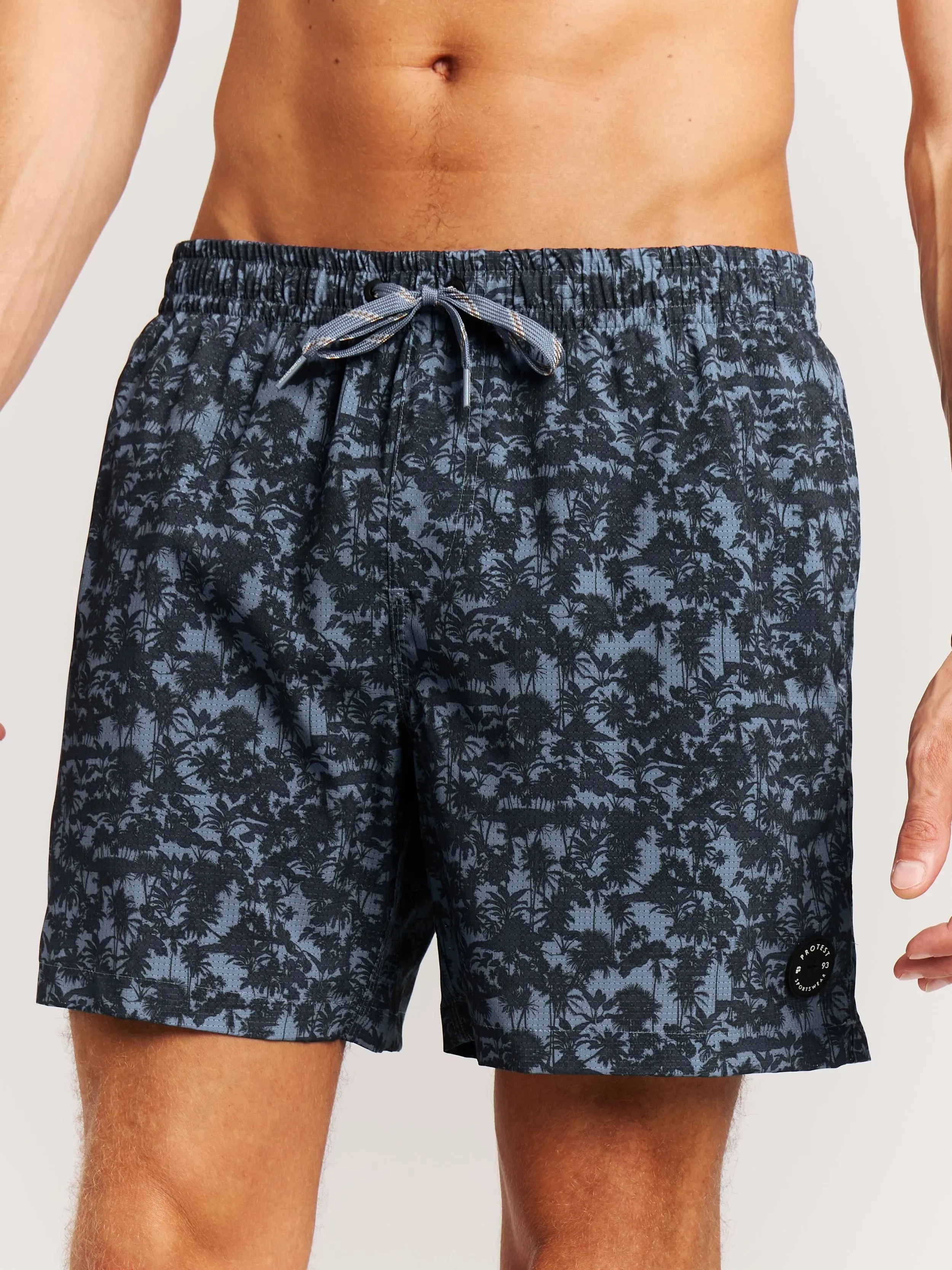 Ein Mann trägt Badehosen mit einem schwarzen Palmenmuster auf blauem Grund. Die Badehose hat eine Kordel am Bund.