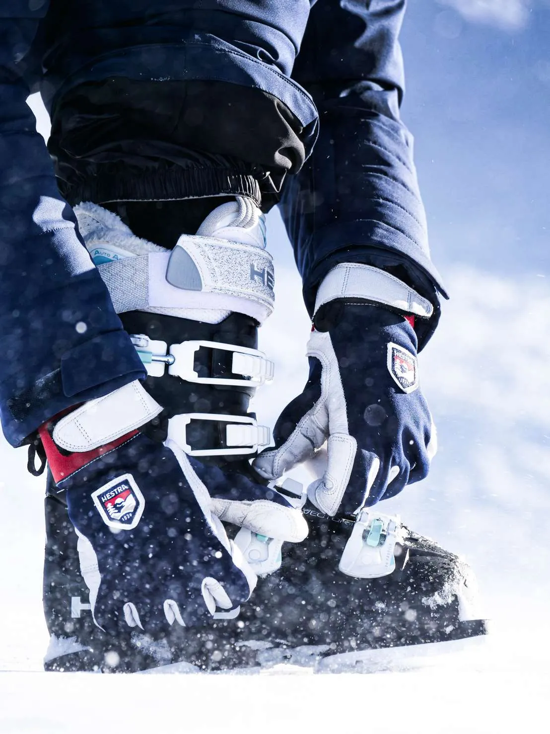 Skifahrer schließt im Schnee mit Hestra-Handschuhen einen Skischuh mit weißen Schnallen.
