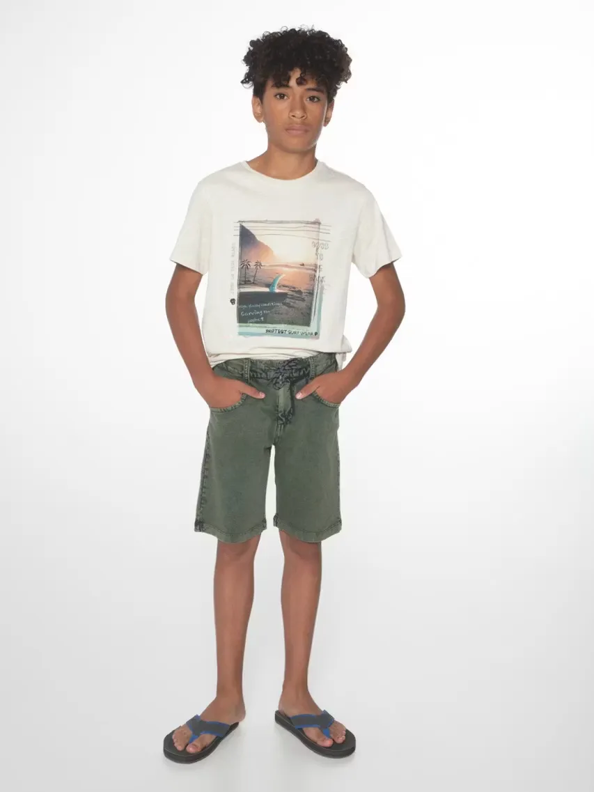 Ein junger Mann mit lockigem Haar steht vor einem weißen Hintergrund. Er trägt ein T-Shirt mit einem Strandmotiv, grüne Shorts und Flip-Flops. Seine Hände stecken in den Hosentaschen.