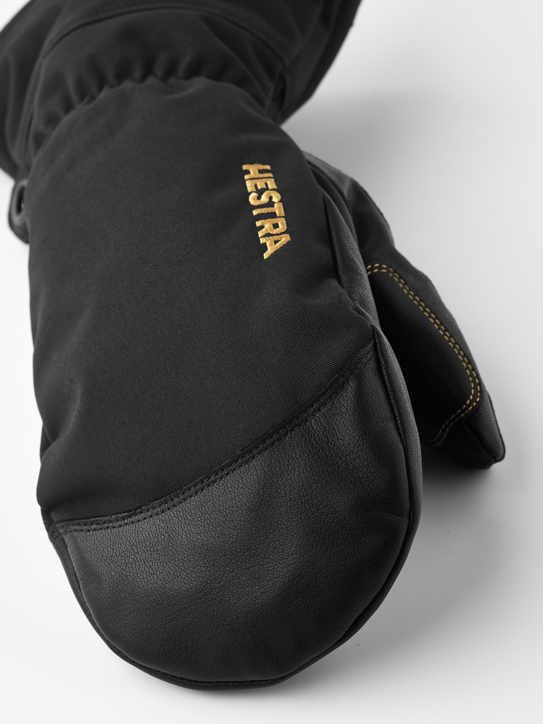 Detailaufnahme eines schwarzen Hestra Fäustlings aus Leder und Stoff, mit dem goldfarbenen Hestra-Logo auf dem Handrücken.