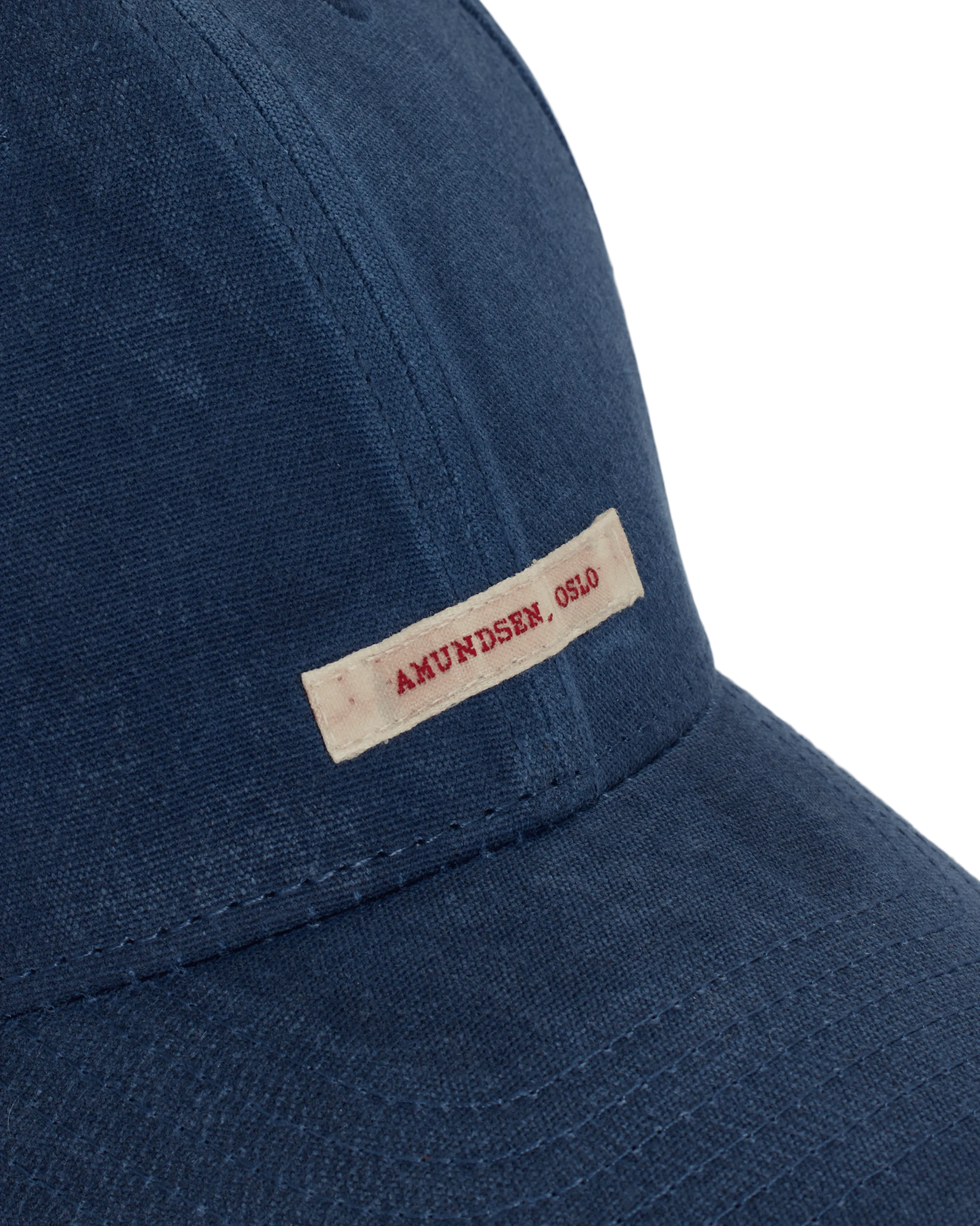 Eine blaue Baseballkappe ist aus einem groben Stoff gefertigt. Ein rechteckiges cremefarbenes Etikett ist an der Seite der Kappe befestigt, mit dem Text „AMUNDSEN, OSLO“ in roter Schrift. Nähte akzentuieren den Schirm und die Seiten der Kappe.