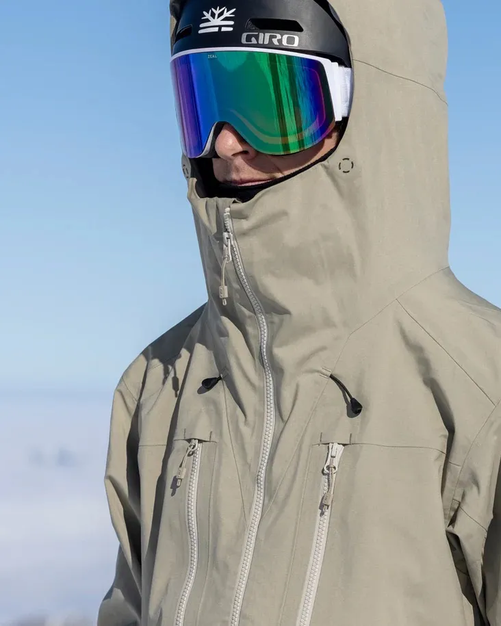 Person in Seneca Rock farbener Z-2 EPE 3L Jacke mit Kapuze, Helm und Skibrille vor blauem Himmel.