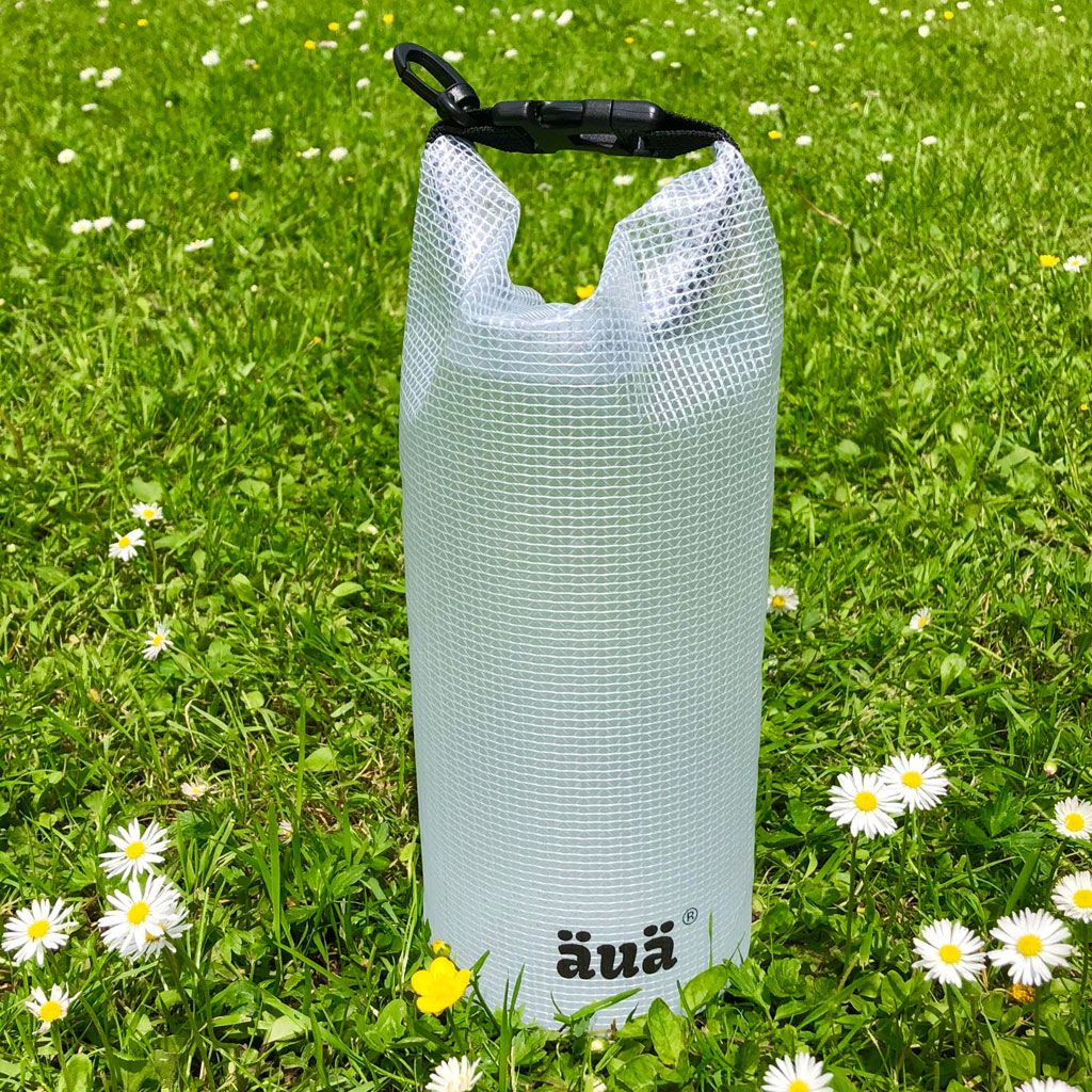 Transparenter Dry Bag auf grünem Gras mit Gänseblümchen, mit schwarzem Verschluss und Logo.