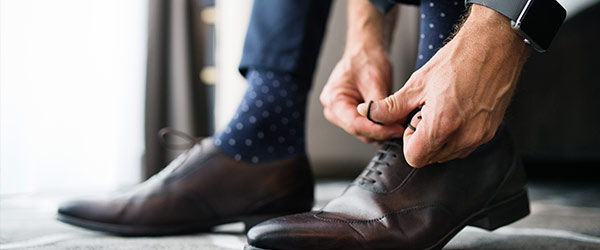 Ein Mann bindet seine braunen Lederschuhe zu, trägt dazu blaue Socken mit weißen Punkten und eine Armbanduhr.