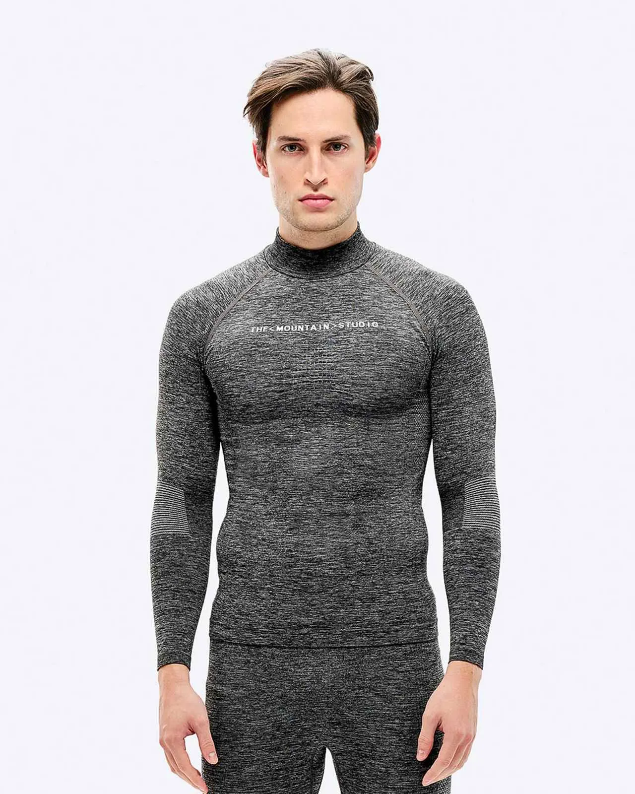 Ein Mann mit braunen Haaren trägt graue Sportbekleidung mit langärmeligem Oberteil und Hose vor einem weißen Hintergrund.
