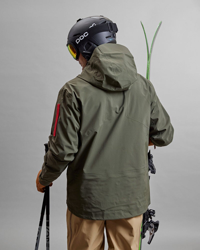 Person von hinten, in grüner Z-2 HD GORE-TEX Pro 3L Shell Jacke, Helm, Skier und Stöcke.
