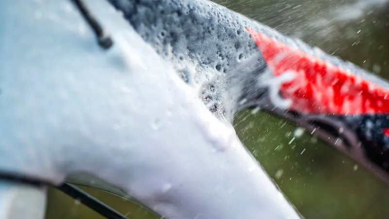 Nahaufnahme: Fahrradrahmen mit Schaum des Fahrradreinigers wird mit Wasser abgespritzt. Rote Farbakzente am Rahmen sichtbar.