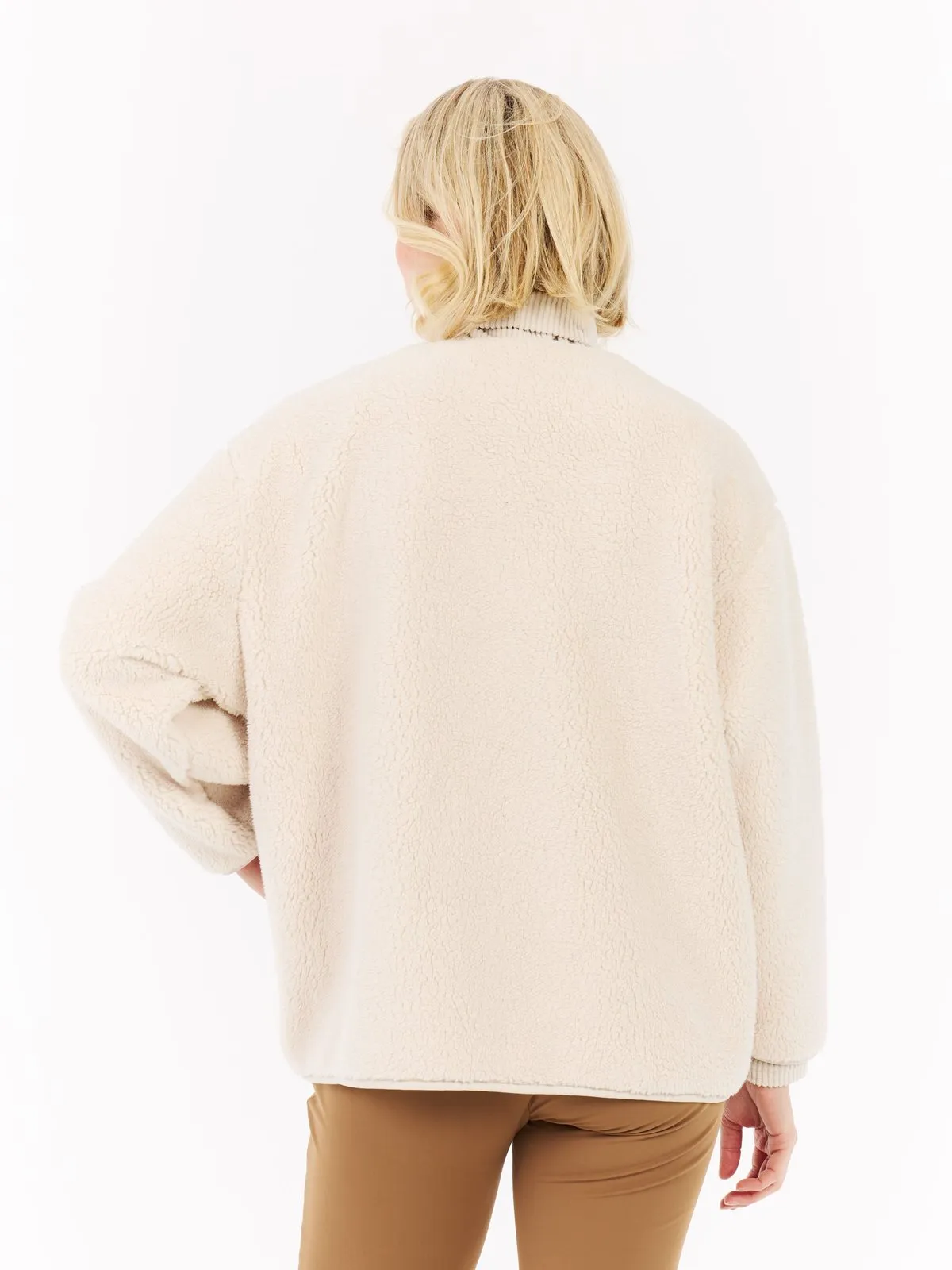 Rückansicht einer Frau mit blondem Haar, die eine cremefarbene Teddyfelljacke und braune Hose vor einem weißen Hintergrund trägt.