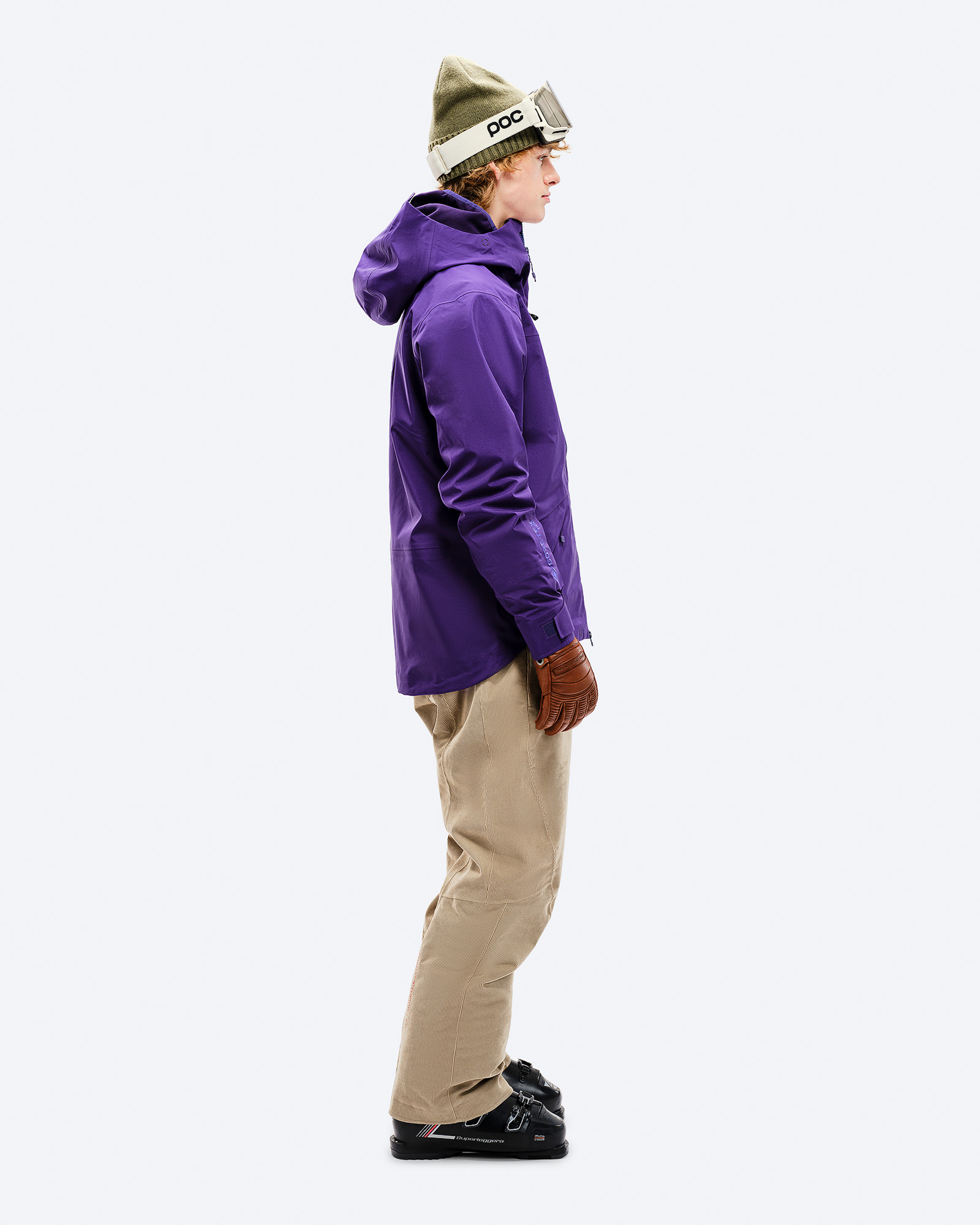 Person von der Seite in lila Z-2 GORE-TEX Jacke, beigen Hosen, Skischuhen, Mütze und Brille vor weißem Hintergrund.