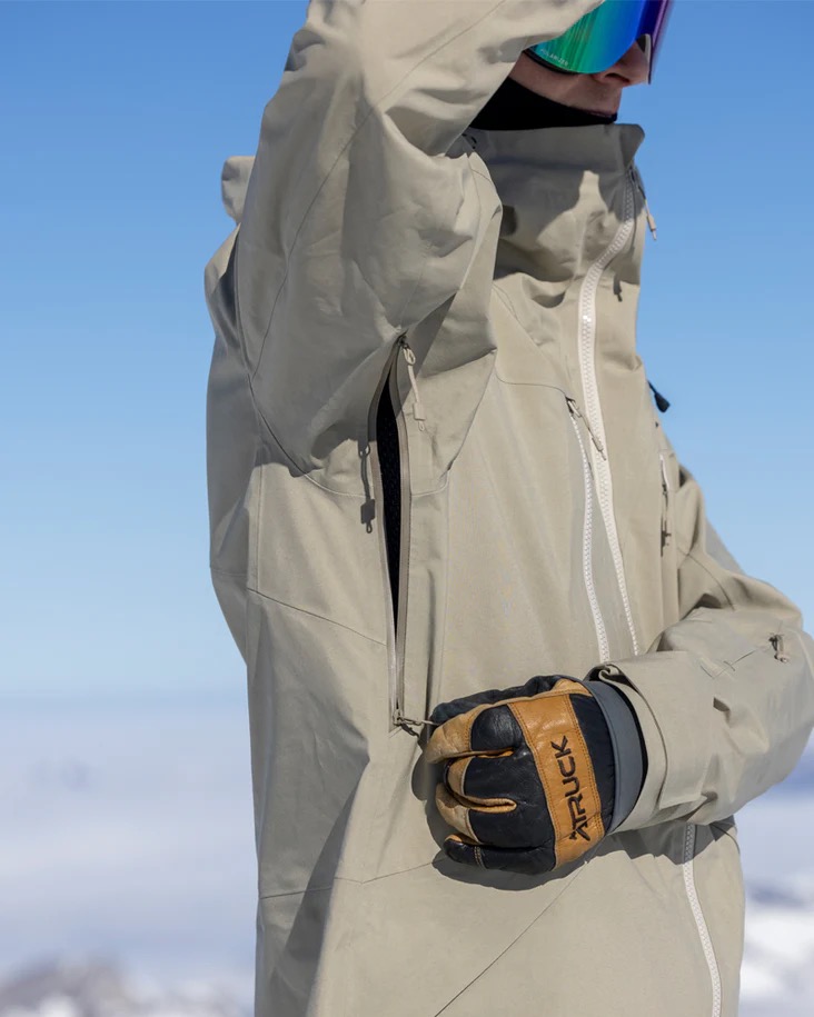Eine Person mit Skibrille und Handschuh, die eine beige Jacke mit Reißverschluss trägt, vor blauem Himmel und verschwommenem Weiß.
