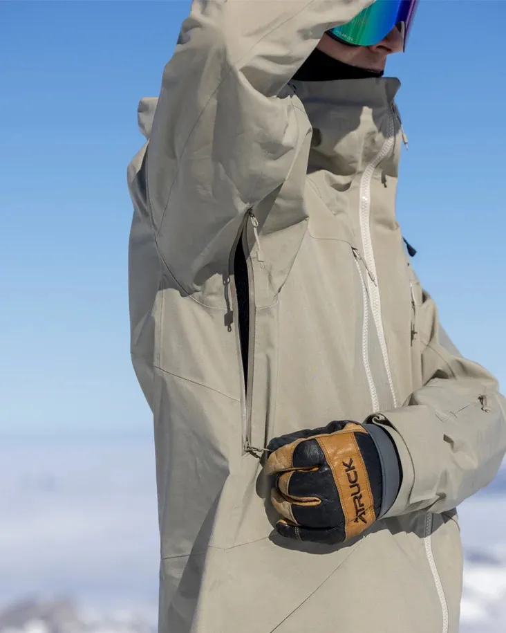 Eine Person mit Skibrille und Handschuh, die eine beige Jacke mit Reißverschluss trägt, vor blauem Himmel und verschwommenem Weiß.