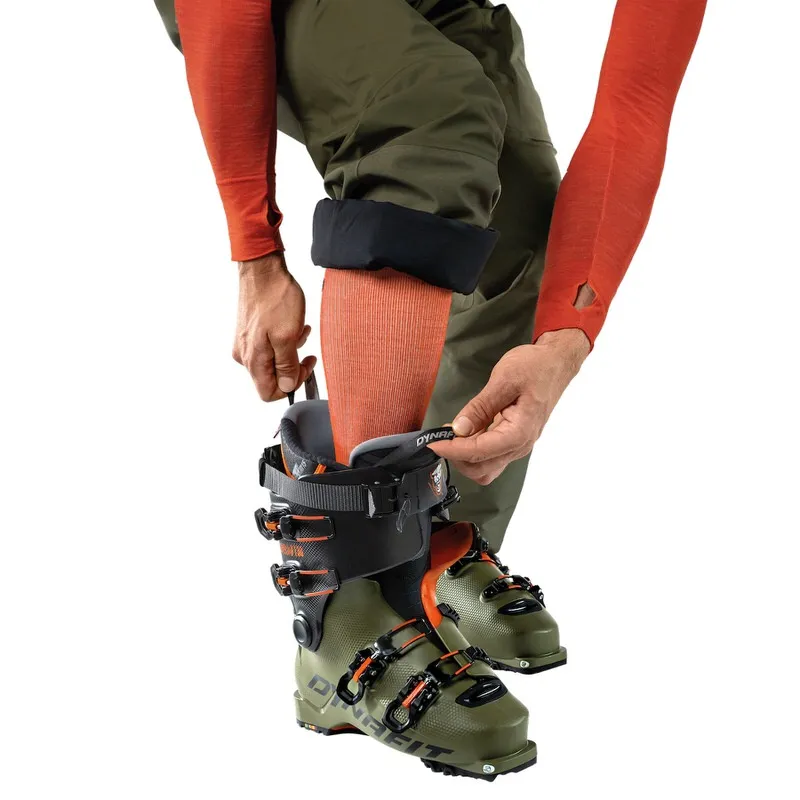 Person zieht Dynafit Tigard Alpine Tech Tourenskischuh an. Der Skischuh ist grün und schwarz mit orangefarbenen Akzenten.