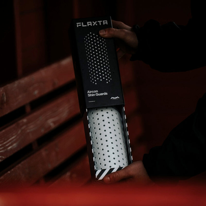 Flaxta Aircon Schienbeinschoner in Verpackung mit Punktmuster, gehalten von Händen.