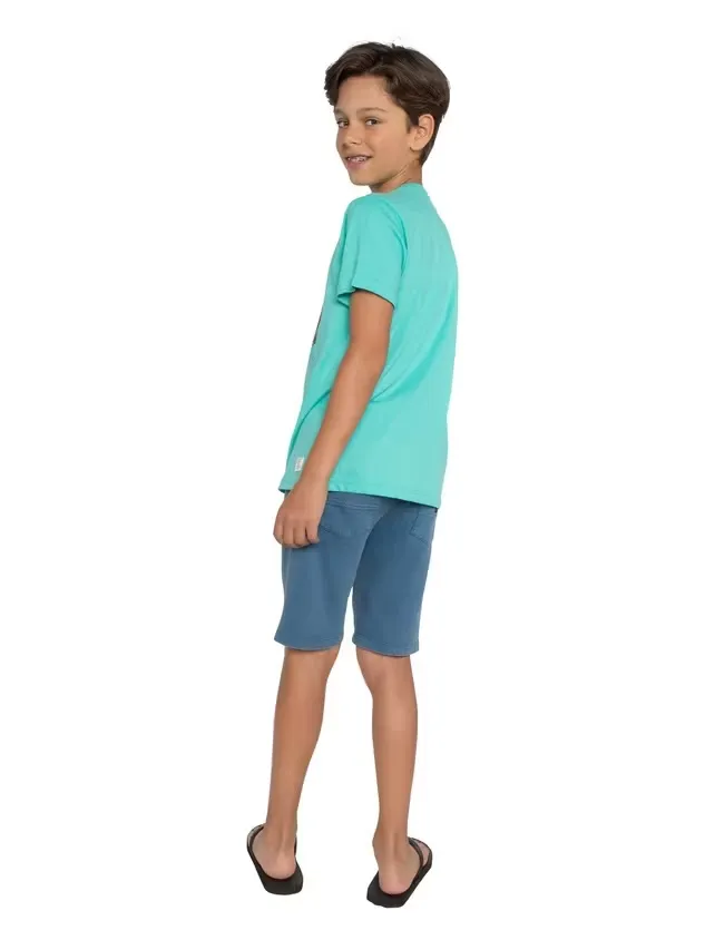 Ein Junge mit dunklem Haar und Zahnspange blickt über seine Schulter in die Kamera. Er trägt ein hellblaues T-Shirt, blaue Shorts und schwarze Flip-Flops. Der Hintergrund ist schlicht weiß.