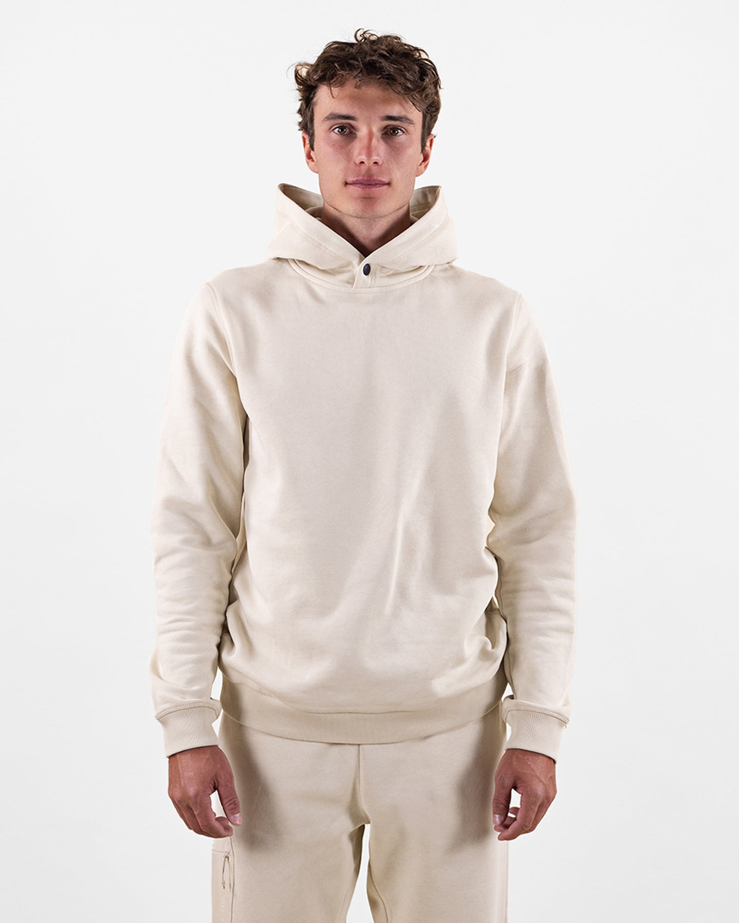 Ein Mann in einem beigen Hoodie und passender Hose vor einem weißen Hintergrund.