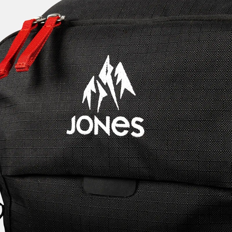 Detailansicht eines schwarzen Jones Further 25L Rucksacks mit weißem Logo und roten Reißverschlüssen.