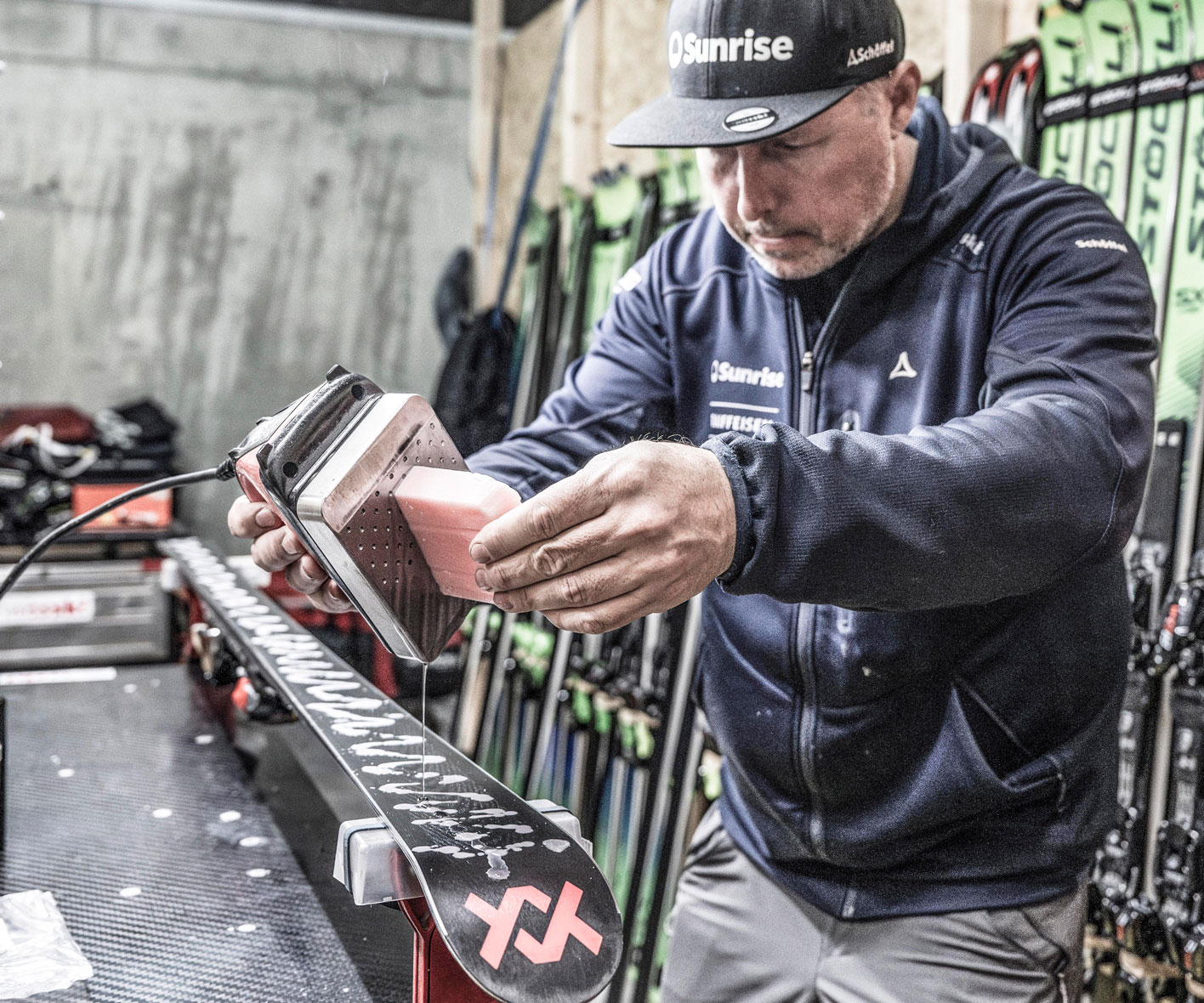FZero Wachs – Das Profiwachs der Swiss-Ski-Athleten jetzt im ski-shop.ch erhältlich Mann wachst einen Ski mit einem Wachseisen und einem rosa Wachsblock, der auf einem Tisch vor einer Skireihe liegt.