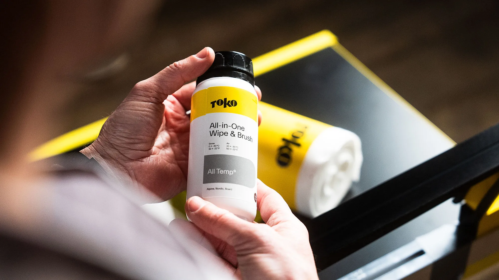 Eine Person hält eine Flasche "Toko All-in-One Wipe & Brush" vor einer Rolle mit Reinigungstüchern. Die Farben sind Gelb, Weiß und Schwarz.
