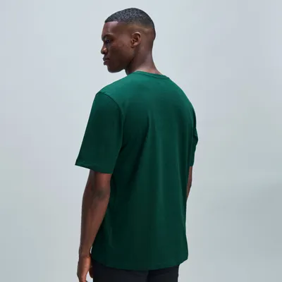 Ein Mann mit kurzem Haar trägt ein grünes T-Shirt und schwarze Hosen, von der Seite aufgenommen.