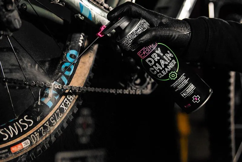 E-Bike Kettenreinigung: Eine Hand sprüht Dry Chain Cleaner auf eine Fahrradkette. Im Fokus: Sprühnebel und die Produktflasche.