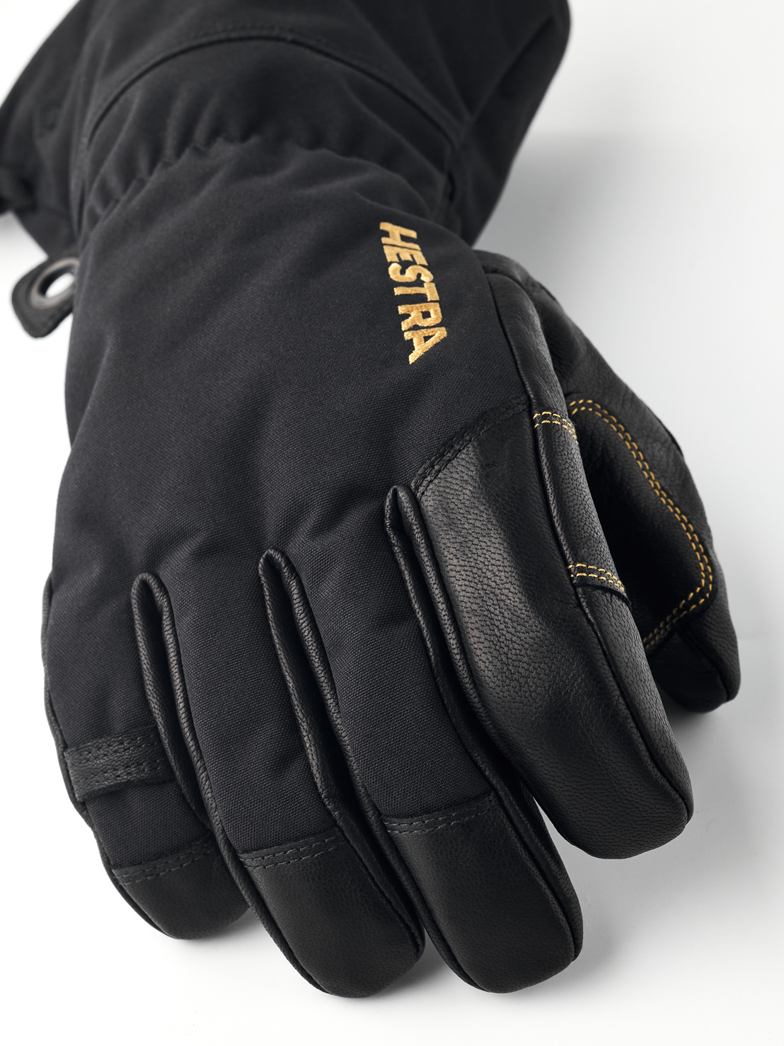 Nahaufnahme eines schwarzen Hestra Gore-Tex Fünffingerhandschuhs mit Lederdetails und goldener Hestra-Aufschrift.
