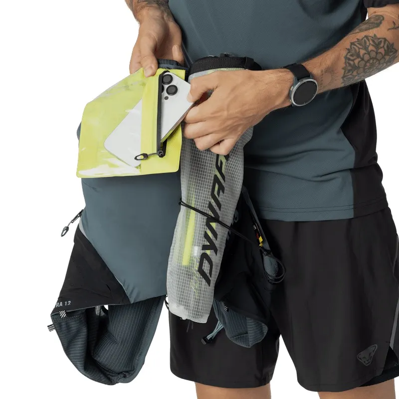 Ein Mann in Sportbekleidung verstaut ein Smartphone in einer gelben, wasserdichten Tasche eines Laufrucksacks. Der Rucksack hat Fächer für Stöcke und Getränke. Er trägt eine Smartwatch am Handgelenk und hat ein Tattoo am Oberarm.