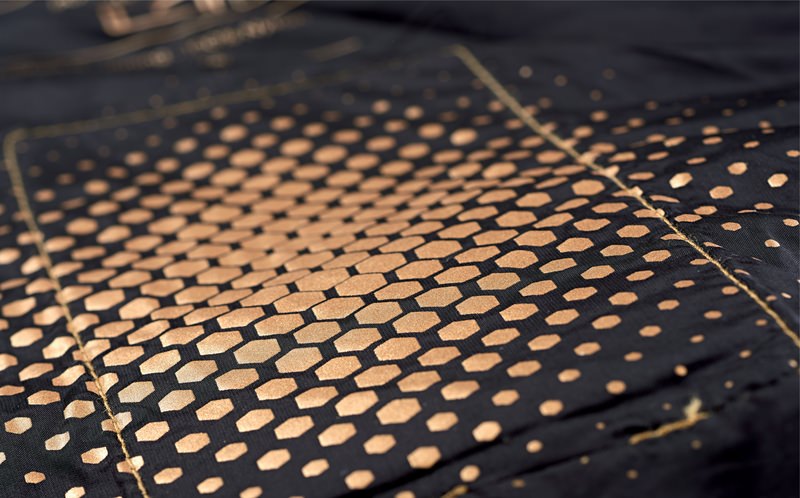 Therm-ic Damen Heizweste Detailaufnahme der Therm-ic Heated Vest Women. Muster aus goldenen Sechsecken auf dunklem Stoff, das in Punkte übergeht.