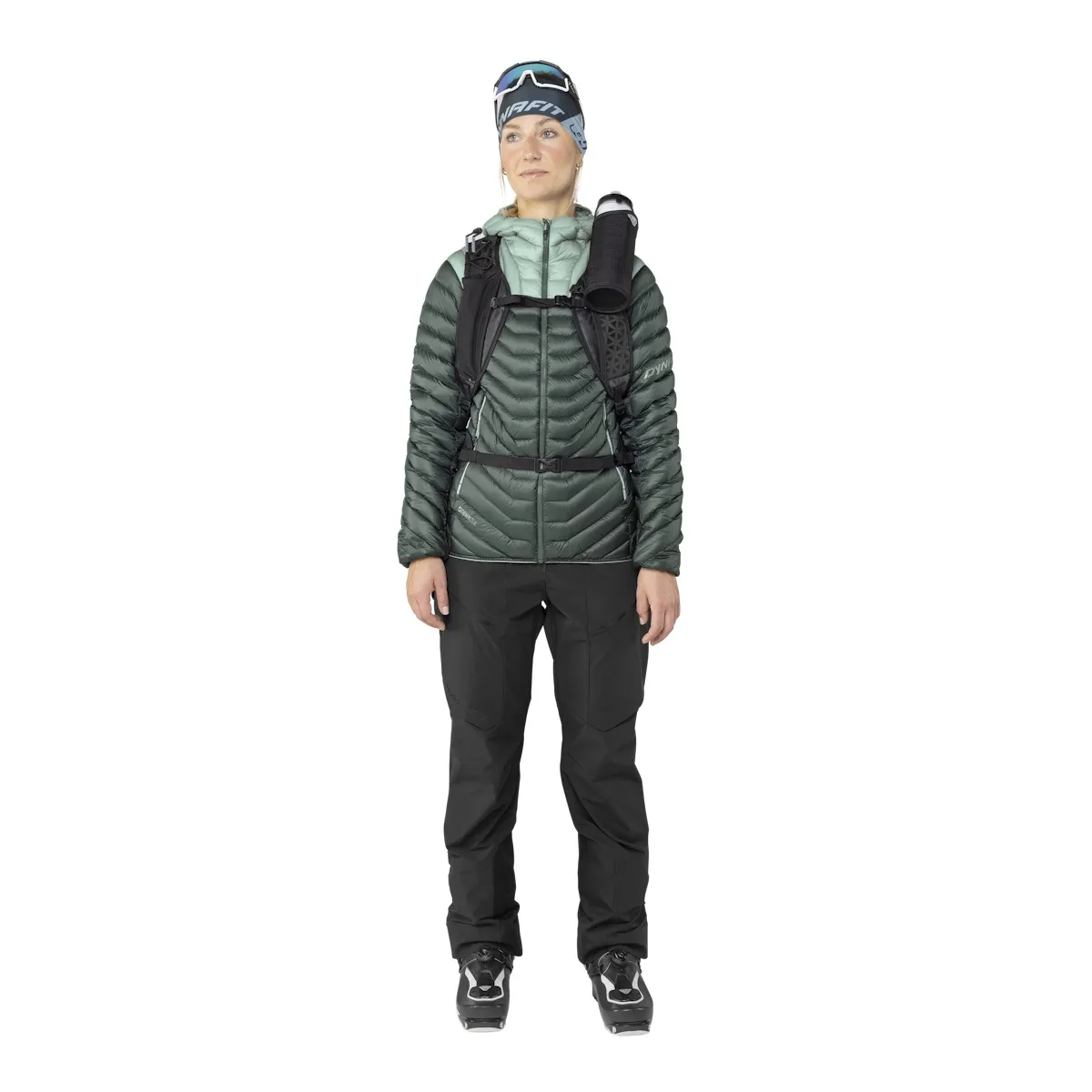 Eine Frau in Ski-Bekleidung, mit Dynafit Ridge Ultralight Daunenjacke in Grün, Hose und Skischuhen.