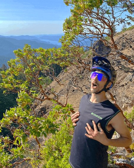 Mann mit Helm und pinker Brille auf einem Berg, Hände auf der Brust, im Hintergrund grüne Bäume und ein See. Afterparty 2000 Feeling.
