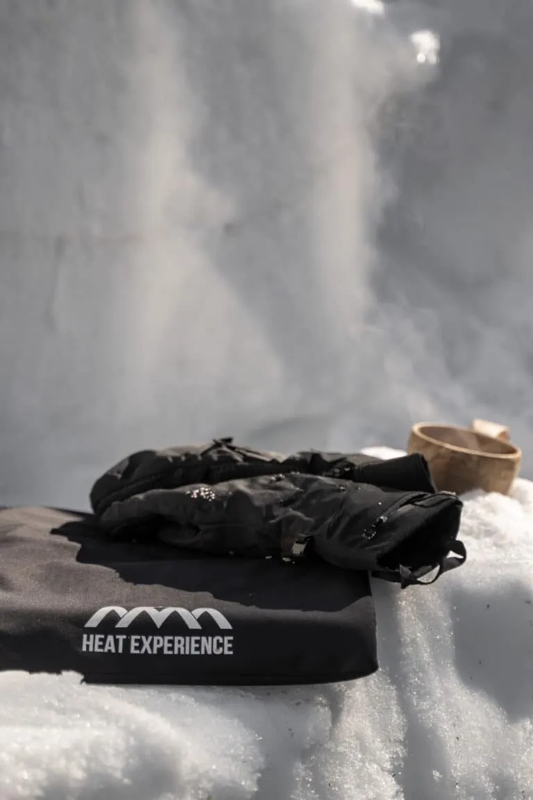 Schwarze Handschuhe mit "Heat Experience"-Logo auf Schnee, eine Holzschale im Hintergrund, Dunst steigt auf.