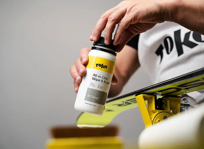 Eine Hand öffnet eine Flasche "TOKO All-in-One Wipe & Brush" über einem Ski. Der Ski liegt auf einer gelben Vorrichtung.