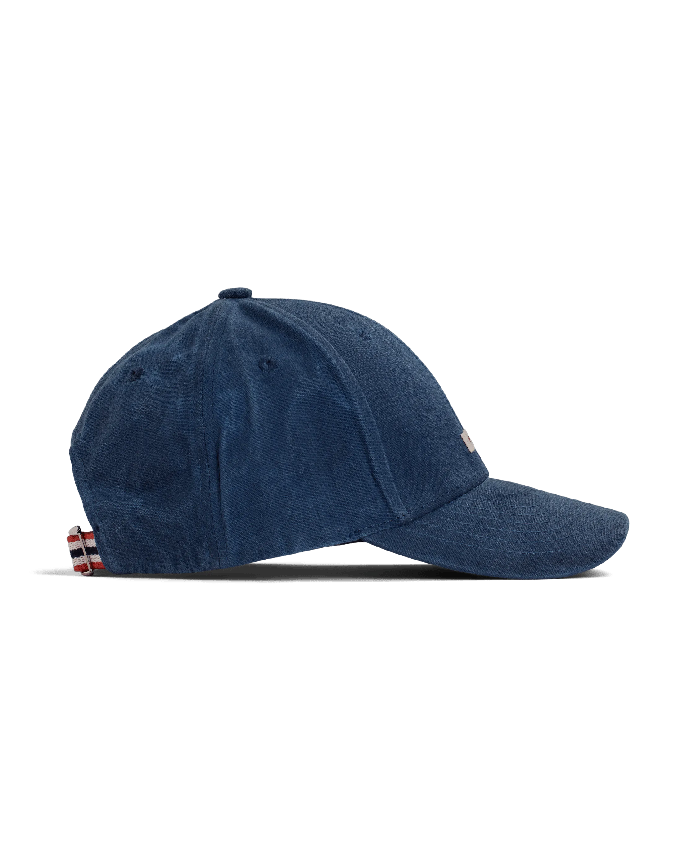 Seitliche Ansicht einer marineblauen Amundsen Waxed Cotton Cap vor einem weißen Hintergrund. Die Kappe hat einen Knopf oben, einen gebogenen Schirm und einen Riemen mit rot-weiß-blauen Streifen am Hinterkopf zur Größenverstellung.