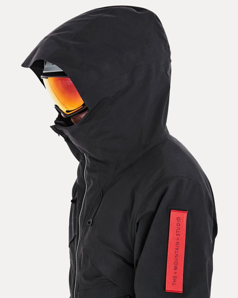 Person mit Skibrille und Kapuze einer schwarzen Gore-Tex 2L Stretch Insulated Jacke. Am Ärmel ein rotes Label.