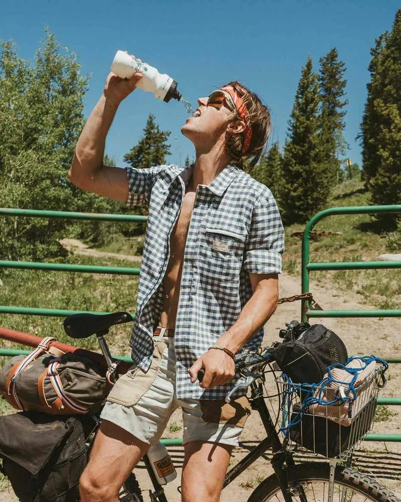 Ein Mann in kariertem Hemd und Shorts trinkt Wasser aus einer Flasche, lehnt an einem Fahrrad vor einer grünen Gittertür.